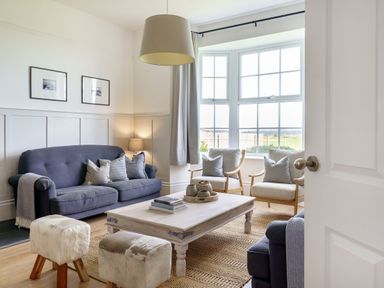 A living room with blue sofas wooden chairs a wooden coffee table and large windows at Ty Carmel in Rhosneigr
