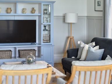 A living room with a blue entertainment unit and television a black sofa with cushions a wooden floor lamp a wooden coffee table and chairs at Ty Carmel in Rhosneigr