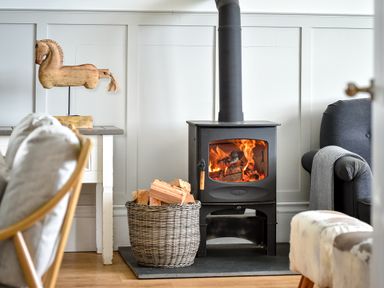 A room with a wood-burning stove next to a basket of firewood a wooden horse sculpture on a table and a gray armchair with a blanket at Ty Carmel in Rhosneigr