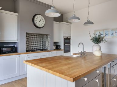 A kitchen with a wooden island countertop white cabinets a wall clock and hanging lights at Ty Carmel in Rhosneigr