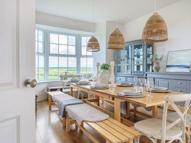 A dining room with a wooden table, benches, chairs, pendant lights, and a large window with countryside view at Ty Carmel in Rhosneigr