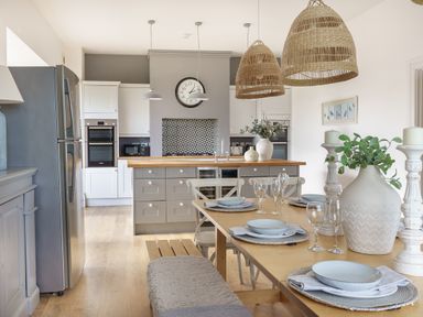 A kitchen and dining area with a wooden table set with plates and glasses and a kitchen island with cabinets and a clock on the wall at Ty Carmel in Rhosneigr