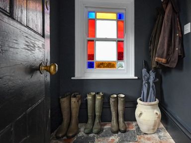 An entryway with mud boots on the floor coats hanging on the wall and umbrellas in a pot under a stained glass window at Ty Carmel in Rhosneigr