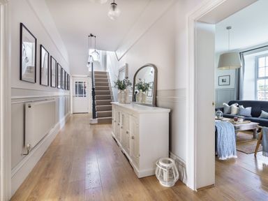 A hallway with framed pictures on the wall a staircase a white sideboard with a mirror and vases with plants and a living room visible through an open door at Ty Carmel in Rhosneigr