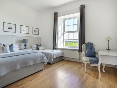 A bedroom with two beds a window with curtains a chair with a pillow and throw and a desk with a lamp at Ty Carmel in Rhosneigr