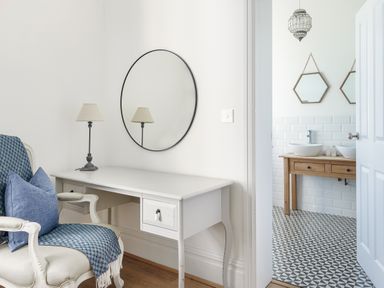 A white desk with a lamp and a round mirror above it next to a chair with a blue throw and cushion with a view into a bathroom with two sinks and hexagon mirrors at Ty Carmel in Rhosneigr