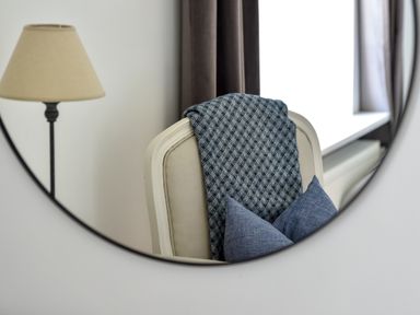 A beige chair with a blue patterned blanket and two blue pillows reflected in a round mirror with a floor lamp nearby at Ty Carmel in Rhosneigr