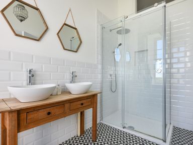 A bathroom with two white vessel sinks on a wooden vanity two hexagonal mirrors and a glass shower enclosure with a rainfall showerhead at Ty Carmel in Rhosneigr