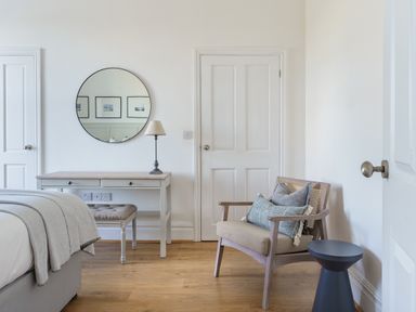 A bedroom corner with a desk, round mirror, lamp, cushioned stool, chair with pillows, and a small side table at Ty Carmel in Rhosneigr