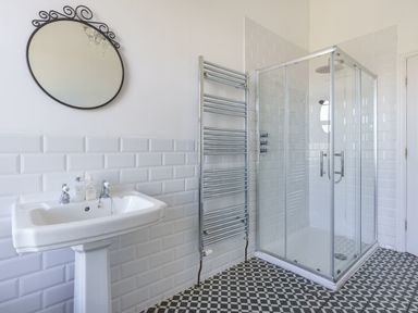 A bathroom with a pedestal sink a round mirror a heated towel rail and a glass shower enclosure at Ty Carmel in Rhosneigr