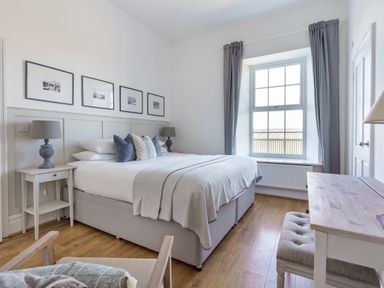 A bedroom with a bed and pillows two bedside tables with lamps a window with curtains and a desk with a bench at Ty Carmel in Rhosneigr