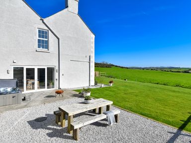A patio area with a wooden picnic table and benches beside a white house with large windows and a green lawn at Ty Carmel in Rhosneigr