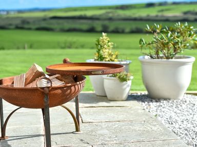 A rusted fire pit with chopped wood on a stone patio with white plant pots and green fields in the background at Ty Carmel in Rhosneigr