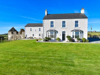 A white two-story house with bay windows and a green lawn under a blue sky at Ty Carmel in Rhosneigr