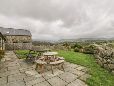 An outdoor patio with two wooden picnic tables near a stone house and a fenced grassy yard with mountains in the background at Y Beudy in Criccieth