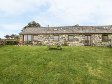 A stone building with a tiled roof and wooden framed windows and doors facing a grassy lawn with picnic tables and a stone bench at Y Beudy in Criccieth