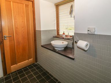 A bathroom with a wooden door a white sink on a dark countertop a window with wooden blinds and a toilet paper holder at Y Beudy in Criccieth