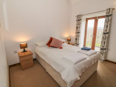 A bedroom with a bed, bedside tables with lamps, curtains, and glass doors at Y Beudy in Criccieth
