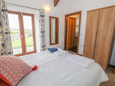 A bedroom with a bed white linens pillows and towels a wooden wardrobe a door leading to another room and glass double doors showing an outdoor area at Y Beudy in Criccieth