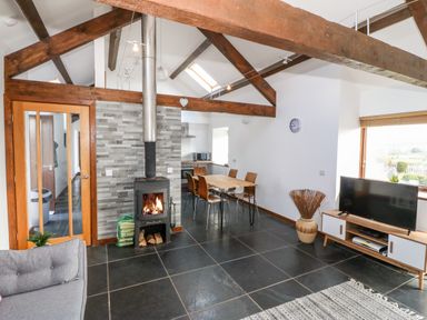 An open living room with exposed wooden beams a wood burning stove a dining table chairs a TV on a stand and a window at Y Beudy in Criccieth