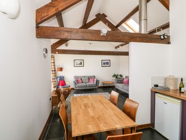 A dining area with wooden table and chairs open to a living room with sofas under exposed wooden beams at Y Beudy in Criccieth