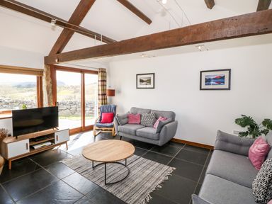 A living room with grey sofas a round wooden coffee table a tv on a stand and large windows at Y Beudy in Criccieth