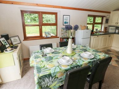 A dining area with a table set for four with green leaf patterned tablecloth and black chairs next to a kitchen with cream cabinets at The Beeches in Narberth