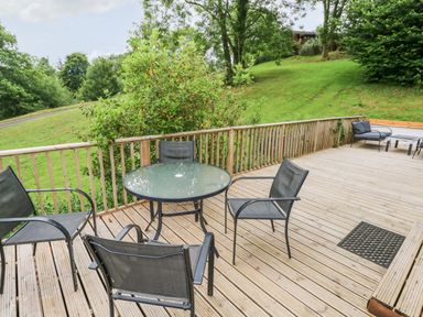 An outdoor wooden deck with a round glass table and four black chairs surrounded by green hills and trees at The Beeches in Narberth