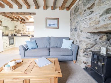 A living room with a grey sofa two cushions a wooden coffee table and a black stove heater by a stone wall at Ysgybor Yd in Chwilog near Criccieth