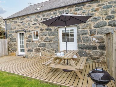An outdoor wooden deck with a picnic table and umbrella next to a stone wall house with two white doors and a barbecue grill on a grass lawn at Ysgybor Yd in Chwilog near Criccieth