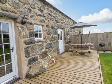 A stone wall with two doors and a window next to a wooden deck with a picnic table umbrella and a barbecue grill at Ysgybor Yd in Chwilog near Criccieth