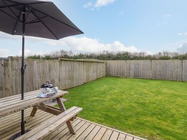 A wooden picnic table with umbrella on a wooden deck next to a grass fenced garden at Ysgybor Yd in Chwilog near Criccieth