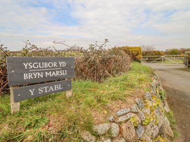 A stone wall with grass beside a road and a metal gate with signs on posts reading Ysgubor Yd Bryn Marsli Y Stabl at Ysgybor Yd in Chwilog near Criccieth
