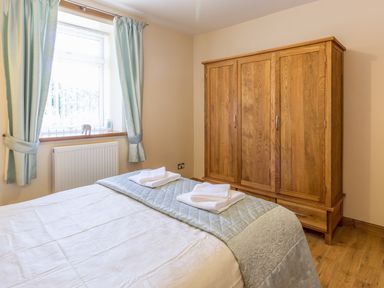 A bedroom with a bed with folded towels on top a wooden wardrobe and a window with light blue curtains at Gardeners Cottage Belford near Bamburgh