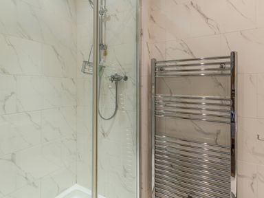 A shower area with glass door and a chrome towel radiator on a tiled wall at Gardeners Cottage Belford near Bamburgh