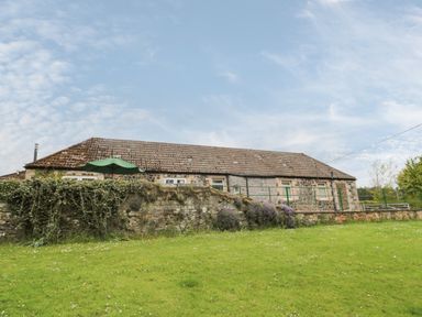 A stone cottage with a tiled roof behind a stone wall and green lawn at Gardeners Cottage Belford near Bamburgh
