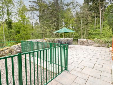 An outdoor patio area with green metal railings a table with chairs and a green umbrella surrounded by trees at Gardeners Cottage in Belford near Bamburgh