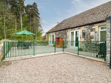 A stone building with a tiled roof and green metal fence around a patio with a table and green umbrella at Gardener's Cottage Belford near Bamburgh
