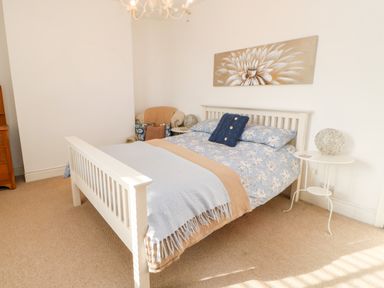 A bedroom with a white bed frame blue and beige bedding a chair with cushions a side table and a flower painting at Lightkeeper House in Amble-by-the-Sea