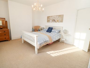 A bedroom with a white bed frame a blue and beige bedspread a wooden dresser a chandelier and a floral wall painting at Lightkeeper House in Amble-by-the-Sea