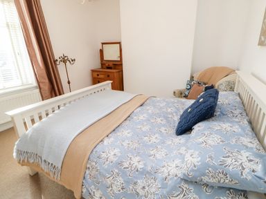 A bedroom with a bed with blue floral bedding a wooden dresser a floral armchair and a window with brown curtains at Lightkeeper House in Amble-by-the-Sea