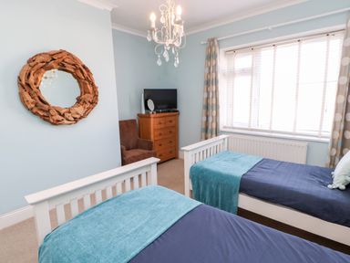 A bedroom with two single beds a wooden dresser a brown armchair a round wooden mirror and a window with curtains at Lightkeeper House in Amble-by-the-Sea