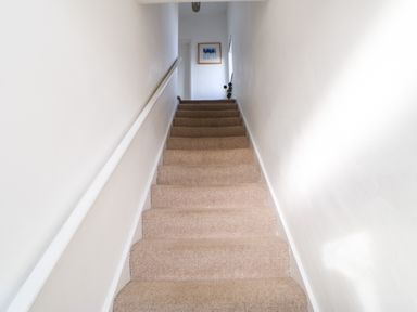 A carpeted staircase with white walls and a handrail leading to a hallway with a framed picture and a small vase of flowers at Lightkeeper House in Amble-by-the-Sea