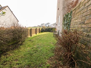 A narrow backyard with grass a bench along a house wall a stone wall on the right and houses in the background at Lightkeeper House in Amble-by-the-Sea