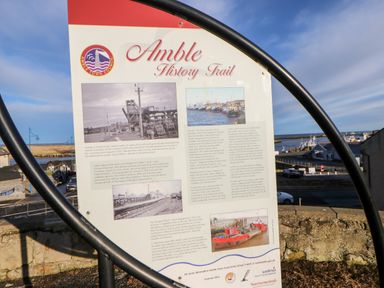 A historical information board about Amble History Trail with images near a harbor in Amble-by-the-Sea