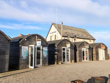 A row of wooden beach huts with glass doors and shops inside with a building labeled coastguard behind at Lightkeeper House in Amble-by-the-Sea