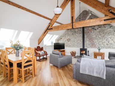 An open living and dining area with a wooden dining table and chairs grey sofas and a wood burning stove under exposed wooden beams at Yr Hen Festri in Y Felinheli
