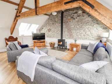 A living room with a gray sofa, armchair, wood burning stove, TV on a wooden stand, and exposed wooden beams at Yr Hen Festri in Y Felinheli