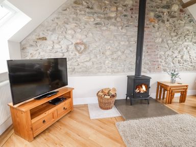 A living room with a stone wall a wood burning stove a basket of firewood a television on a wooden stand and nested wooden tables at Yr Hen Festri in Y Felinheli