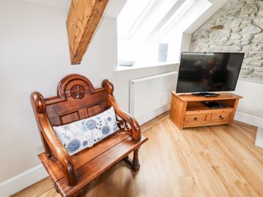 A wooden bench with a floral cushion and a television on a wooden stand in a room with a stone wall at Yr Hen Festri in Y Felinheli